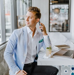 A photo of a man on his phone, sitting at a cafe with a laptop in front of him